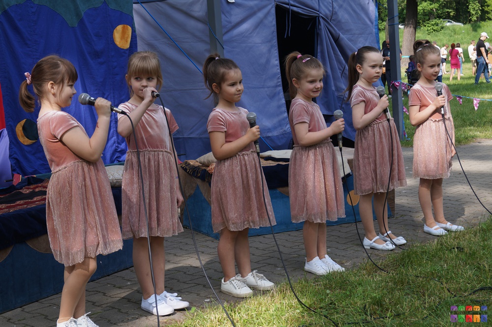 Zdjęcie przedstawiające 6 dziewczynek stojących w rzędzie i trzymających mikrofon w ręce. W tle widać rozłożony niebieski namiot oraz chodnik i trawnik.