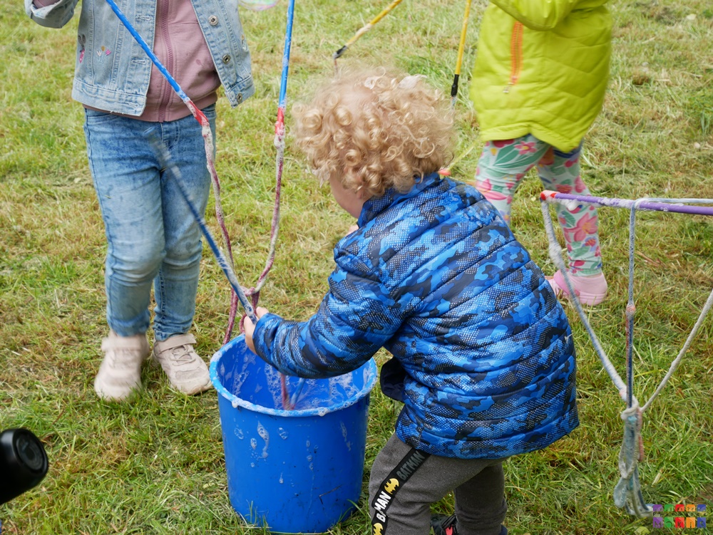 Zdjęcie przedstawiające dziecko zanurzające sznurki w wiaderku. Najprawdopodobniej będzie robić duże Bańki mydlane. W tle trawnik.