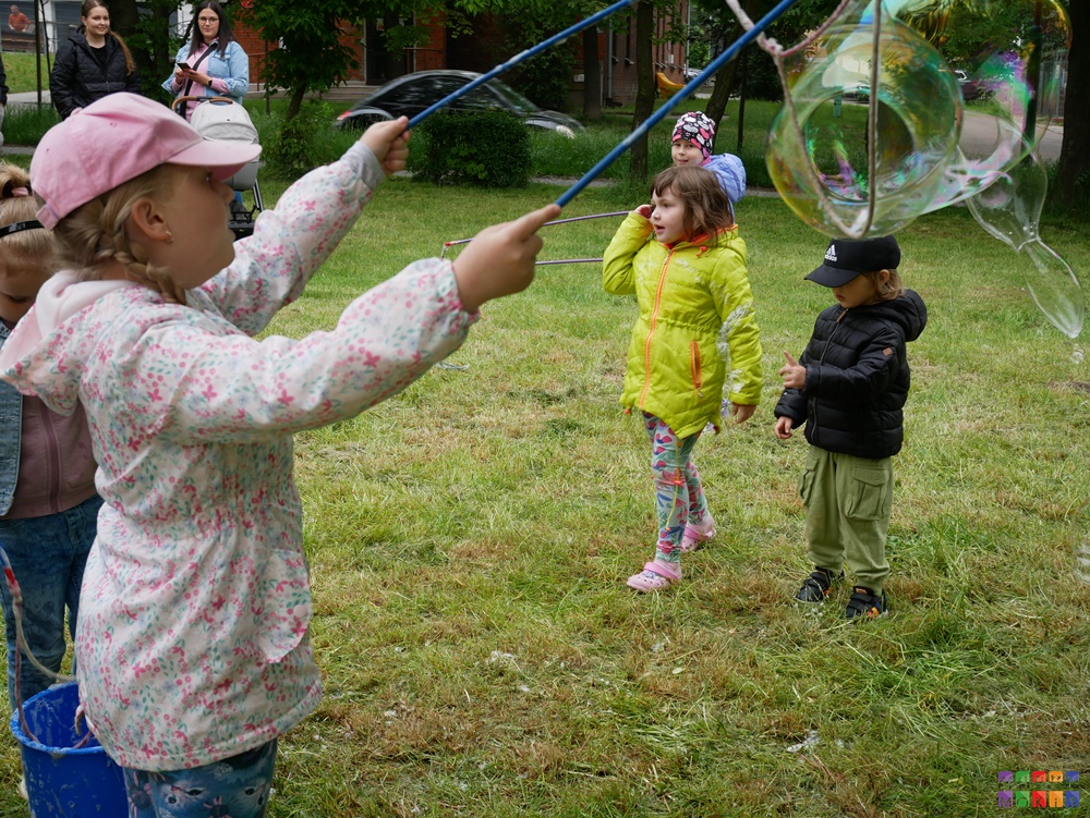 Dziecko stojące na trawniku i robiące bańki mydlane z sznurków.. W tle widać inne dzieci bawiące się.