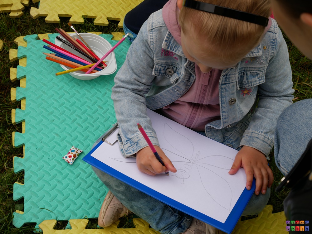 Zdjęcie przedstawiające siedzące dziecko, dziewczynka na piankowej macie i rysujące obrazek na białej kartce papieru