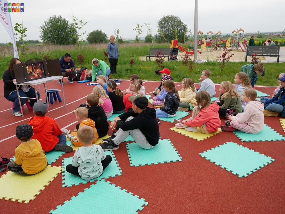 Zdjęcie przedstawiające grupę dzieci siedzących tyłem na boisku. W tle widać rozstawiony teatrzyk na stole oraz powiewające na wietrze flagi i plac zabaw.