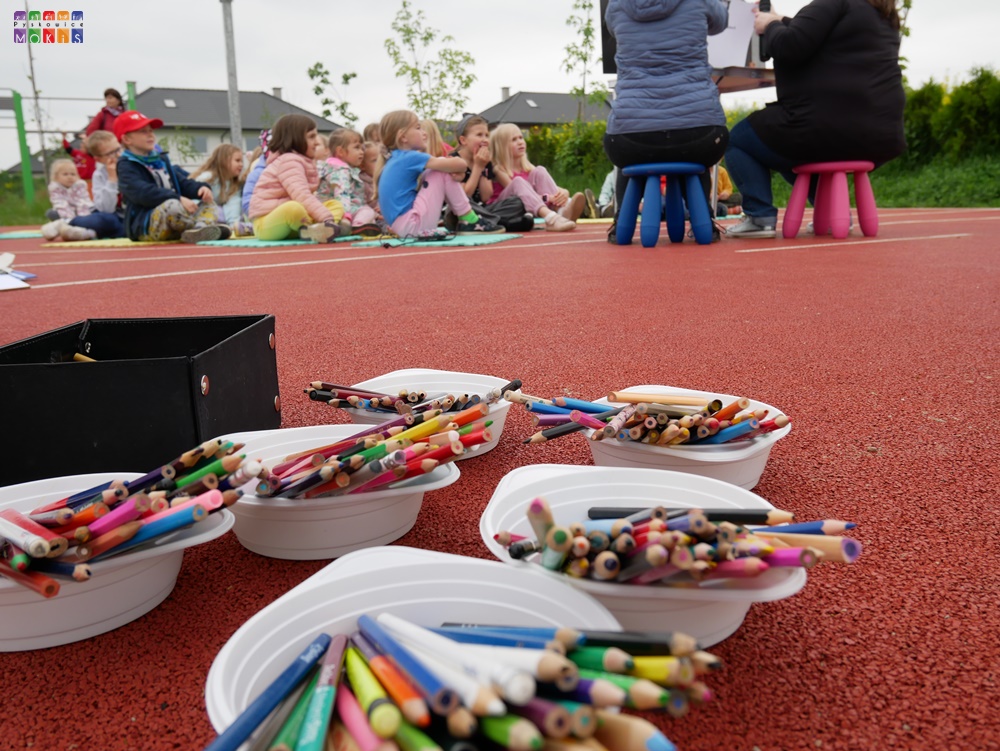 Zdjęcie przedstawiające rozstawione na boisku miseczki z kolorowymi kredkami. W tle siedzące dzieci.