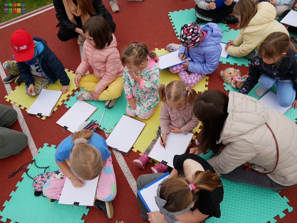 Zdjęcie przedstawiające siedzące dzieci na piankowych, kolorowych matach i rysujących coś na kartkach papieru.