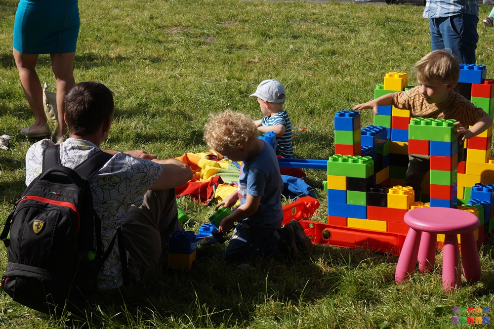 Zdjęcie przedstawiające bawiące się dzieci na trawniku. Układają coś z dużych, kolorowych klocków