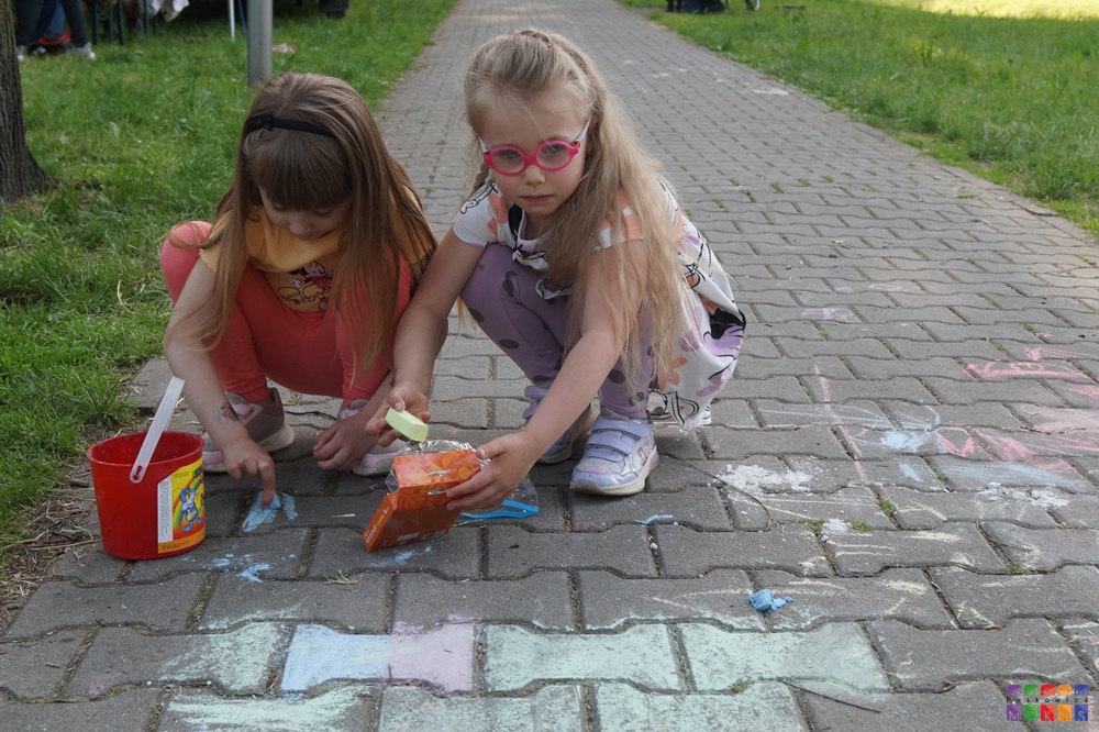 Zdjęcie przedstawiające dwie dziewczynki przysiadające na chodniku oraz rysujące kredą po nim.
