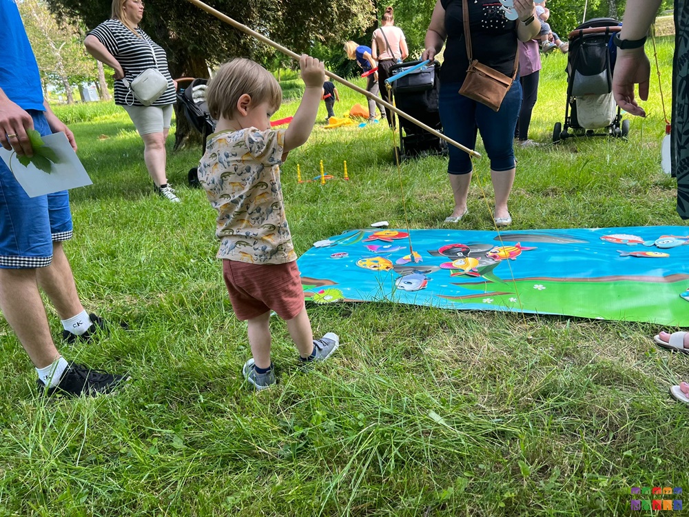 Zdjęcie przedstawiające bawiącego chłopca stojącego na trawniku i trzymającego kij. W tle rozłożona duża mata do gier.