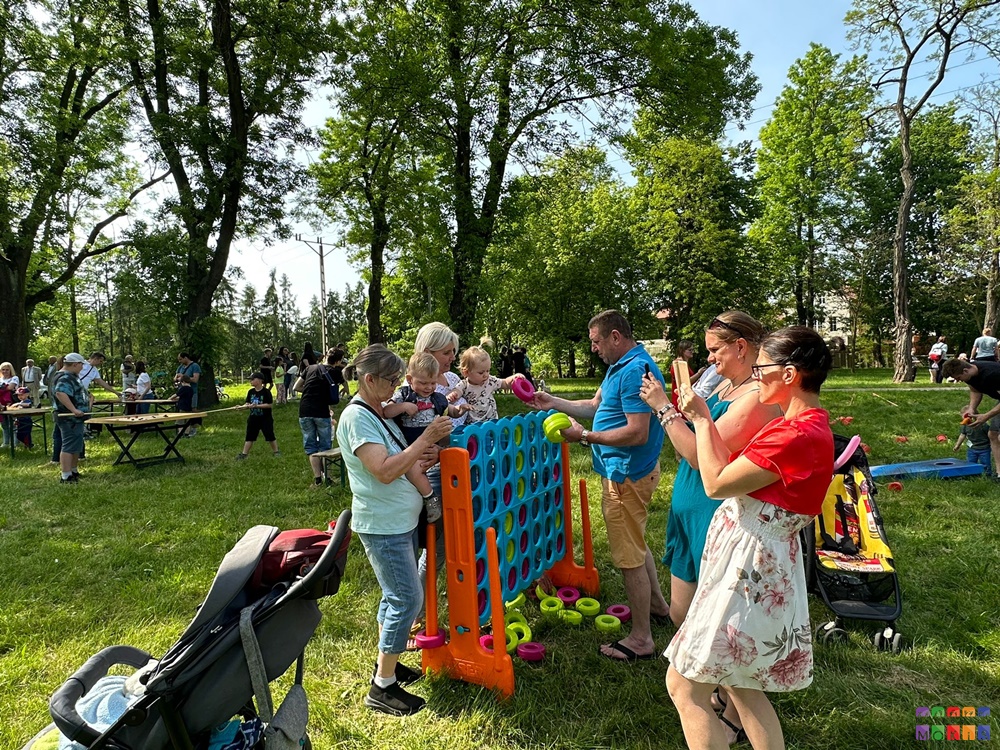 Zdjęcie przedstawiające grupę ludzi bawiących się w grę stojącą pomiędzy nimi. Jest to niebieska ściana z otworami. W tle drzewa parkowe.
