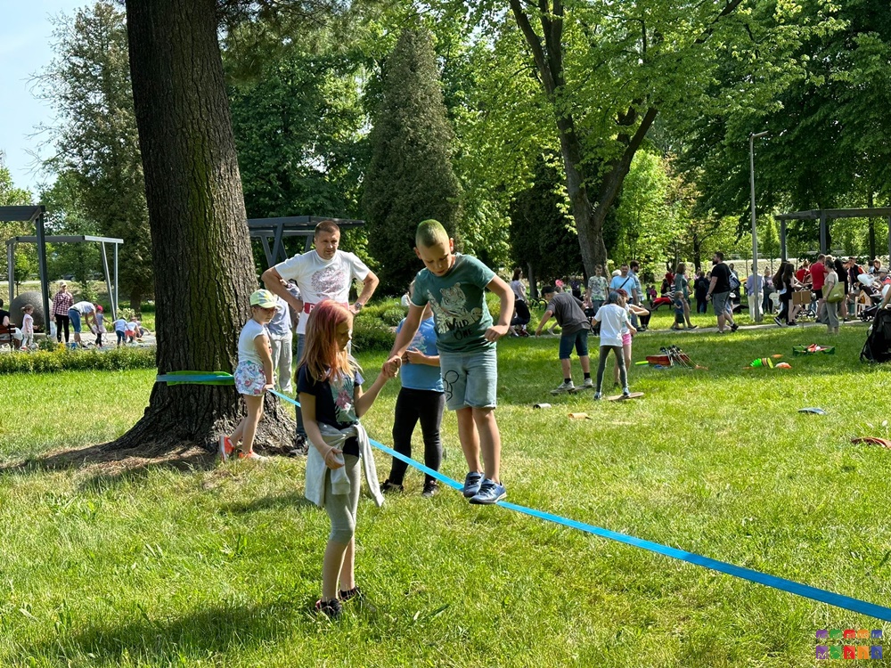 Na zdjęciu znajduje się chłopak chodzący po linie. Dziewczyna trzyma chłopaka za rękę. W tle widać grupy ludzi, trawnik, drzewa parkowe.