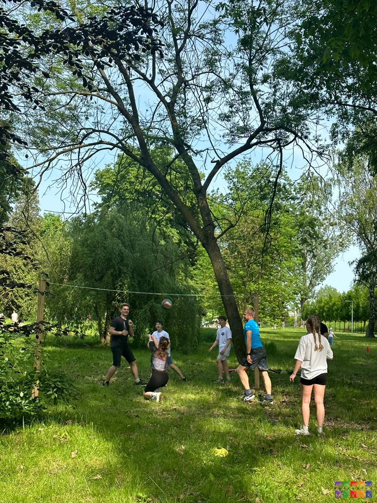 Na zdjęciu grupa młodzieży grającą w siatkówkę. W tle trawnik oraz drzewa parkowe