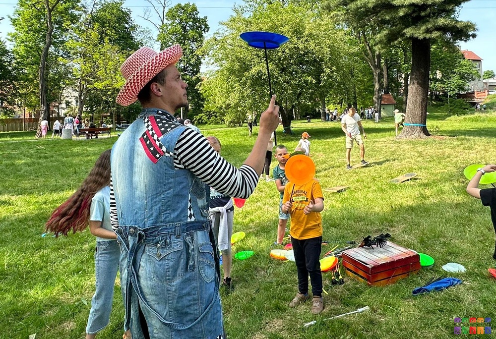 Zdjęcie przedstawiające mężczyznę kręconego talerz na patyku. w tle bawiące się dzieci, trawnik oraz drzewa parkowe.