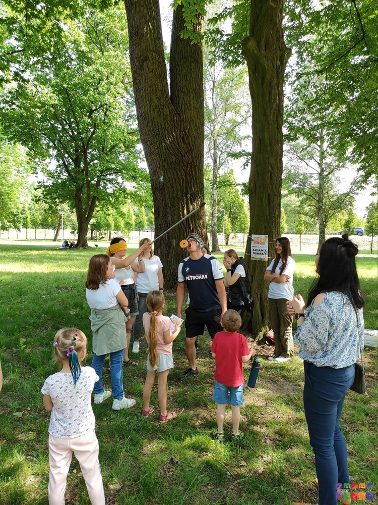 Zdjęcie przedstawiające grupę ludzi na tle pieni z drzew jedna z nich tak jak by jadła pączka zawieszonego na wędce.