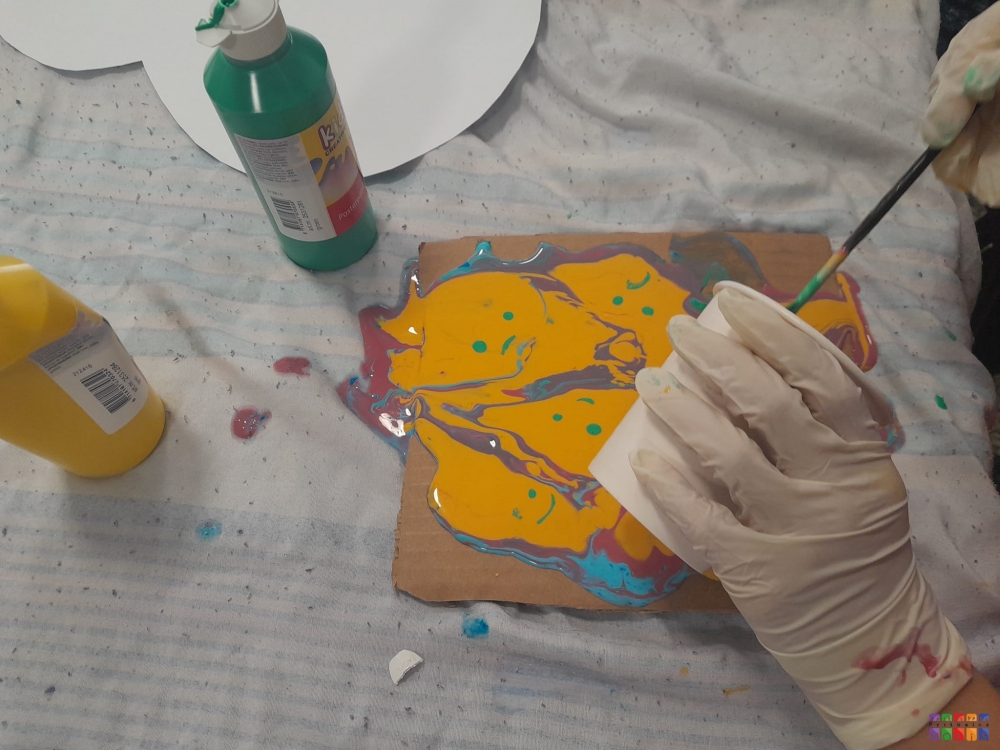 Zdjęcie przedstawiające stół z kawałkiem dykty, na którą ktoś rozlewa farbę z kubeczka. Obok stoją butelki z farbami.