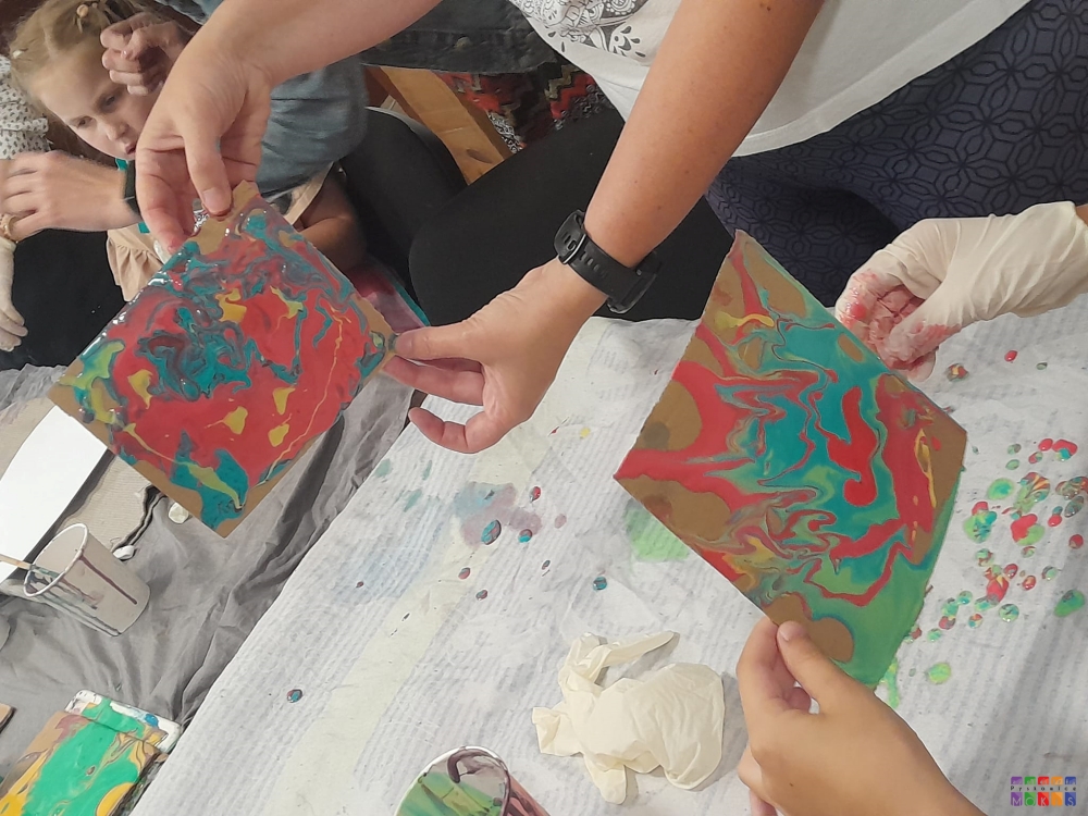 Zdjęcie przedstawia trzymające w rękach dykty nad stołem z namalowanymi obrazami z farb które były na nich rozlane