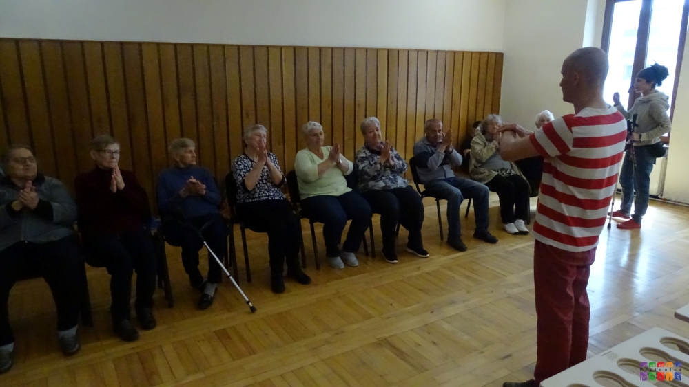 Na zdjęciu grupa siedzących seniorów ze złożonymi dłońmi. Przed nimi stojący mężczyzna i pokazujący ćwiczenia jakie mają robić.