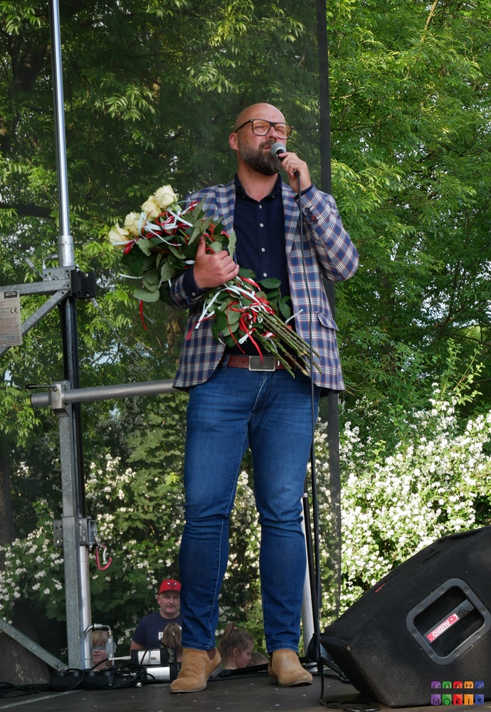Zdjęcie przedstawiające stojącego mężczyznę z bukietem róż w ręku i mówiącego coś do mikrofonu