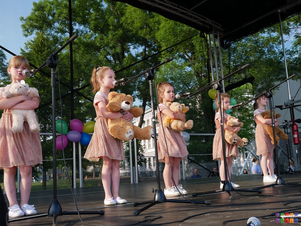Zdjęcie przedstawiające śpiewające dzieci do mikrofonów na statywach. W rękach trzymają pluszowe misie.
