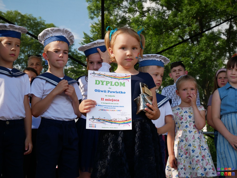 Zdjęcie przedstawiające stojącą dziewczynkę z dyplomem i statuetką w ręku. Za nią stoją inne dzieci.
