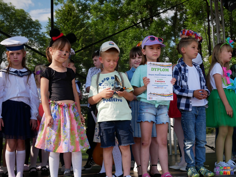 Zdjęcie przedstawiające stojące dzieci z dyplomem i statuetką w ręku. Za nią stoją inne dzieci.