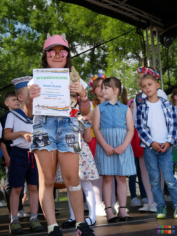 Zdjęcie przedstawiające stojącą dziewczynkę z dyplomem i statuetką w ręku. Za nią stoją inne dzieci.