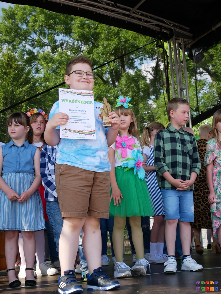 Zdjęcie przedstawiające stojącego chłopaka z dyplomem i statuetką w ręku. Za nim stoją inne dzieci.