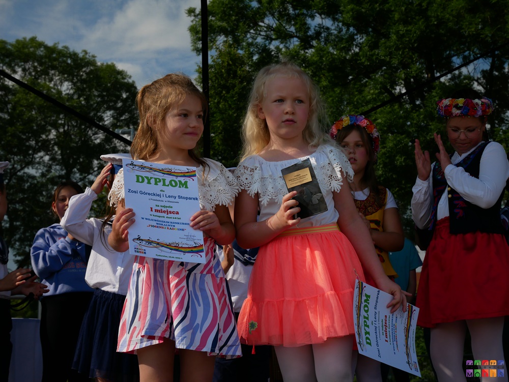 Zdjęcie przedstawiające stojące dwie dziewczynki z dyplomem i statuetką w ręku. Za nią stoją inne dzieci.