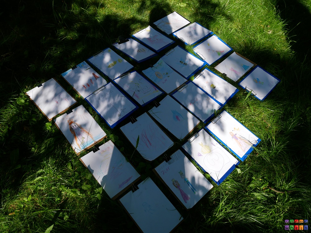 Zdjęcie przedstawia rozłożone kartki z rysunkami na trawniku.