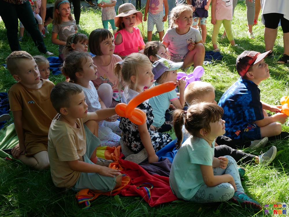 Zdjęcie przedstawia siedzące dzieci na trawniku, niektóre z nich ma balony w rękach.