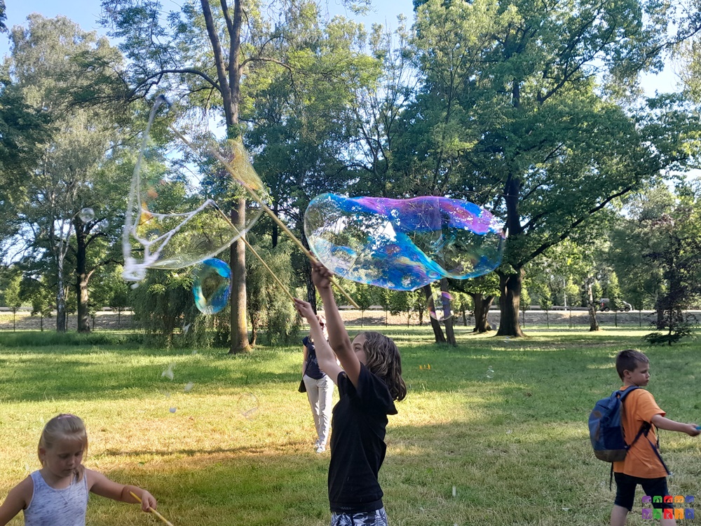 Zdjęcie przedstawiające bawiące się dzieci w parku. Na pierwszym planie dziecko robi ogromną bańkę mydlaną przelatującą nad jego głową. W tle trawnik oraz drzewa parkowe.