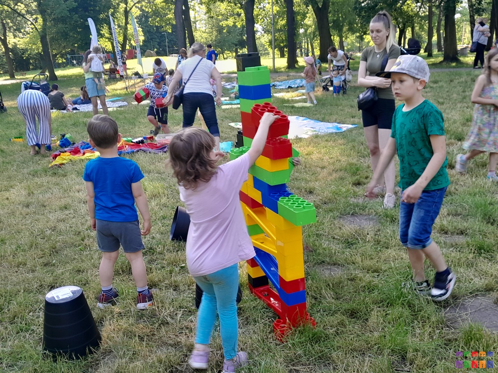 Zdjęcie przedstawiające bawiące się dzieci w parku. na pierwszym planie dziecko układa coś z dużych kolorowych klocków. W tle drzewa parkowe, trawnik.