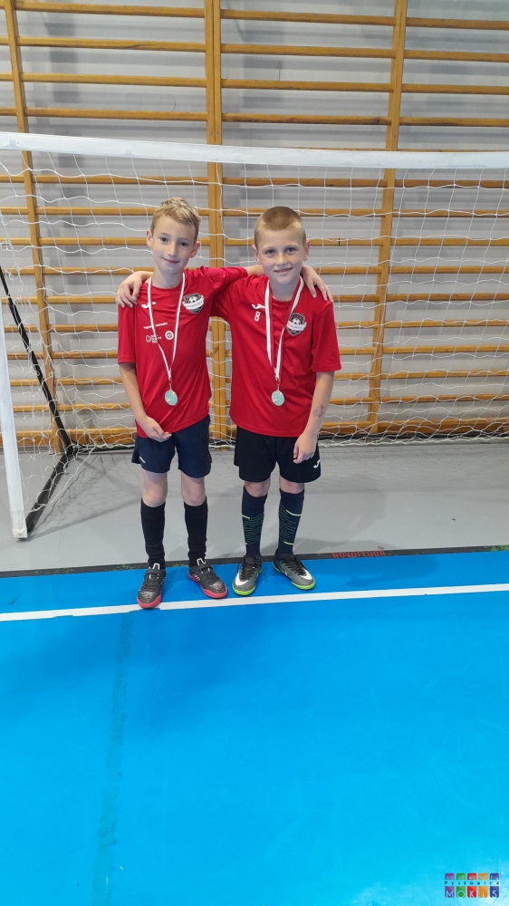 Zdjęcie przedstawiające dwoje chłopców z medalami na szyi. W tle bramka piłkarska na hali sportowej