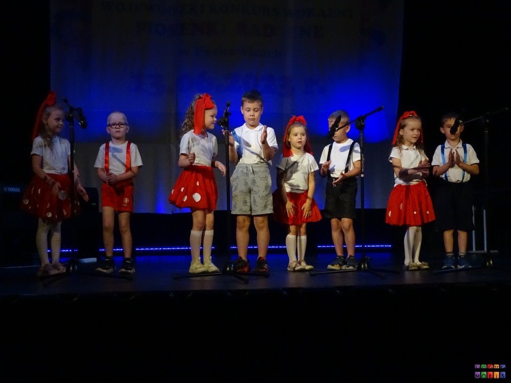 Zdjęcie przedstawiające grupę dzieci śpiewających znajdujących się na scenie przed mikrofonami na statywach. Ubrani kolorowo.