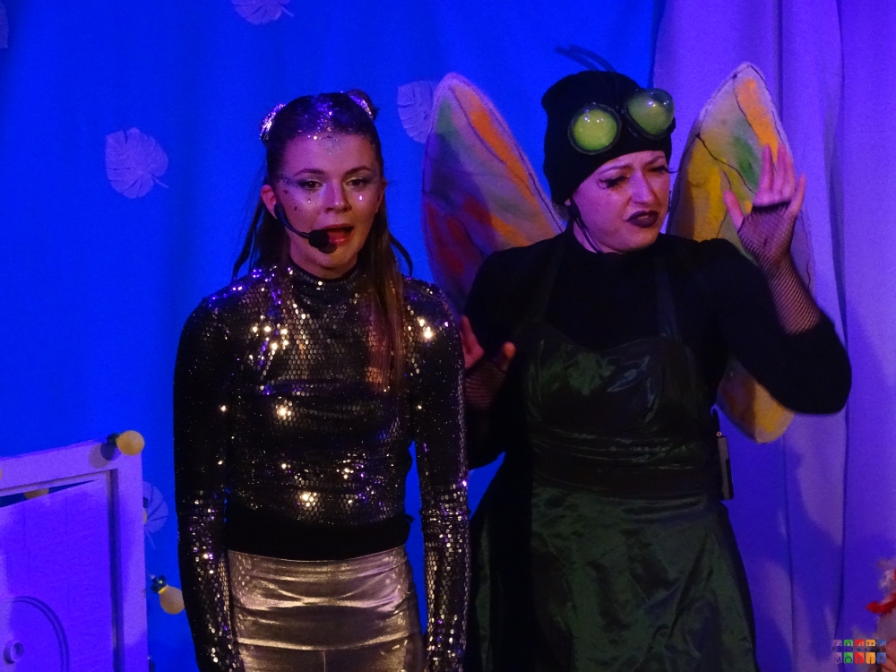 zdjęcie przedstawiające grę dwóch aktorek stojących na scenie mokis jedna po prawej przebrana za muchę a druga z lewej za iskrę w stroju błyszczącym srebrnym