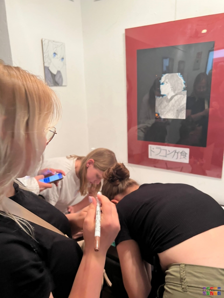Zdjęcie przedstawiające kobiety podpisujące coś . Są one obrócone bokiem do fotografa. W tle na ścianie wiszące obrazy.