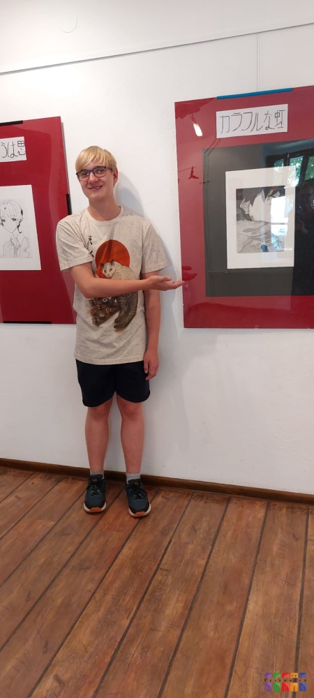 Zdjęcie przedstawia chłopca stojącego przy ścianie i wyciągniętą prawą rękę z kierunku lewym pokazującym wiszący obraz na ścianie.