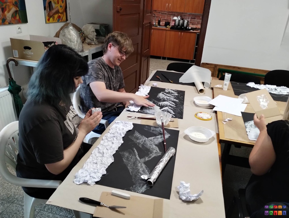 Zdjęcie przedstawia grupę ludzi siedzących i stojących przy stole. Wykonują pracę metodą faktury na kartkach papieru. Stół zapełniony różnymi farbami, pędzelkami.