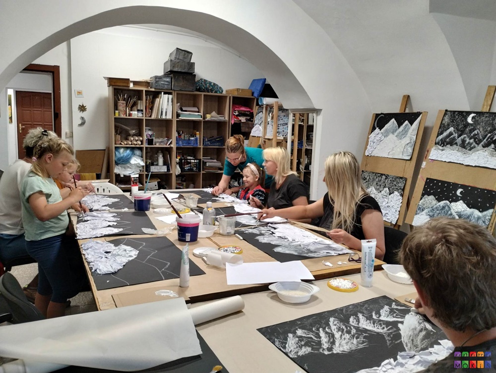 Zdjęcie przedstawia grupę ludzi siedzących i stojących przy stole. Wykonują pracę metodą faktury na kartkach papieru. Stół zapełniony różnymi farbami, pędzelkami.
