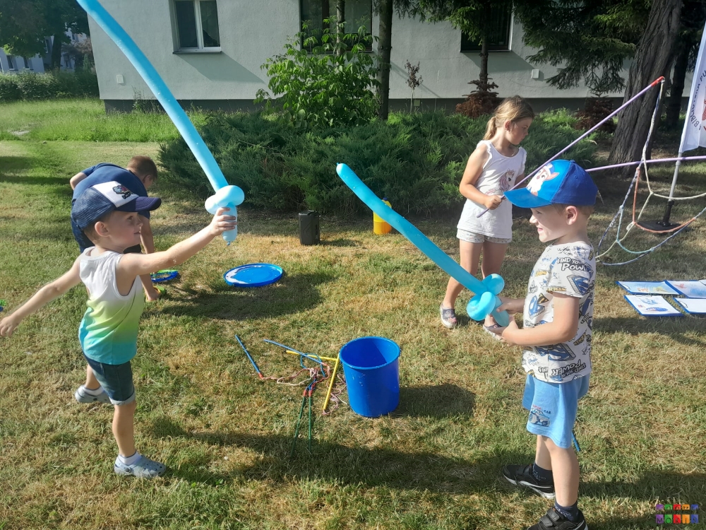 Zdjęcie przedstawiające bawiące się dzieci na trawniku. Na pierwszym planie dzieci z mieczami zrobionymi z balonów.