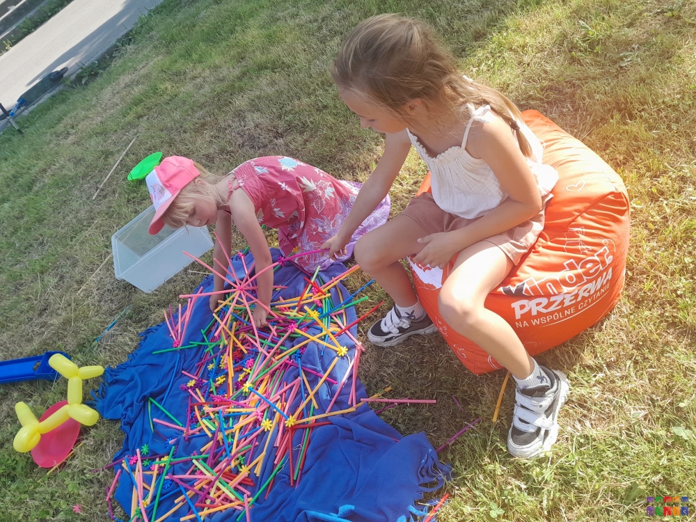 Zdjęcie przedstawiające bawiące się patyczkami kolorowymi dwie dziewczynki na trawniku. jedna po prawej stronie siedzi na pufie w kształcie sześcianu, druga klęczy na trawniku.