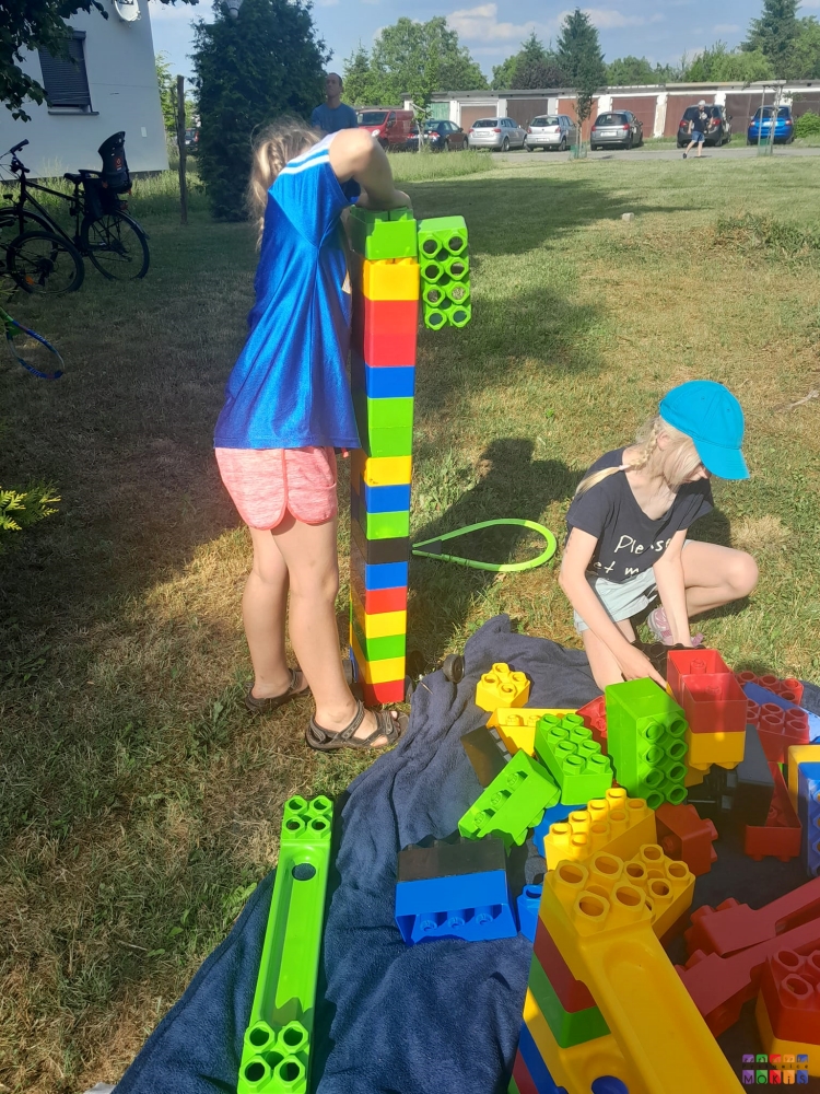Zdjęcie przedstawiające bawiące się dzieci dużymi kolorowymi klockami. Po lewej dziewczyna składa dużą wieżę. W tle widać zaparkowane w rzędzie samochody.