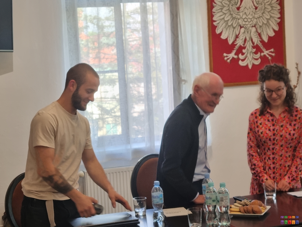 Zdjęcie przedstawiające 3 osoby dwóch mężczyzn jedna kobieta stojących przy stole na którym znajduje się talerz z ciastkami i woda butelkowana. W tle widać okno oraz powieszone na ścianie godło Polski.