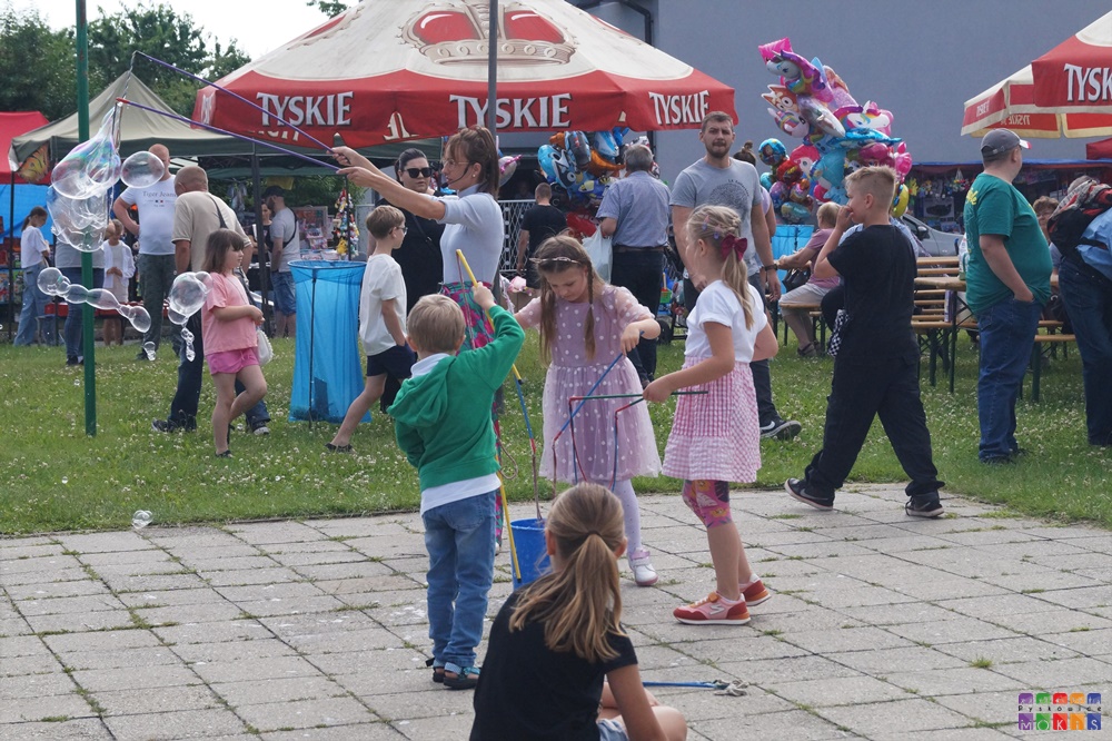 Zdjęcie przedstawia bawiące się dzieci w trakcie festynu. W tle widać parasole oraz stoły plenerowe na których siedzą ludzie.