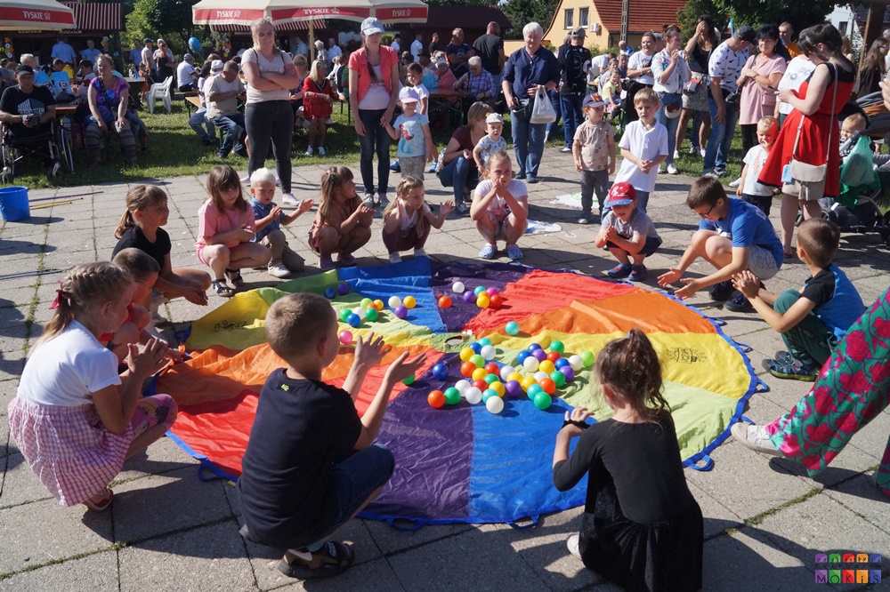 Zdjęcie przedstawia bawiące się dzieci z chustą na której leżą piłeczki w trakcie festynu. W tle widać parasole oraz stoły plenerowe na których siedzą ludzie.