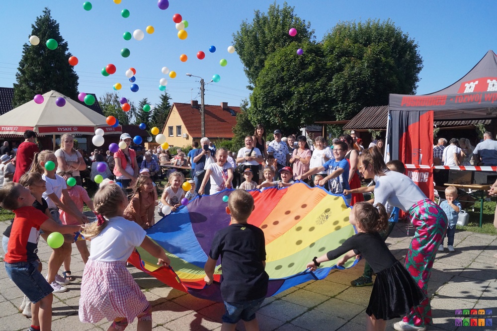 Zdjęcie przedstawia bawiące się dzieci w trakcie festynu. W tle widać parasole oraz stoły plenerowe na których siedzą ludzie.