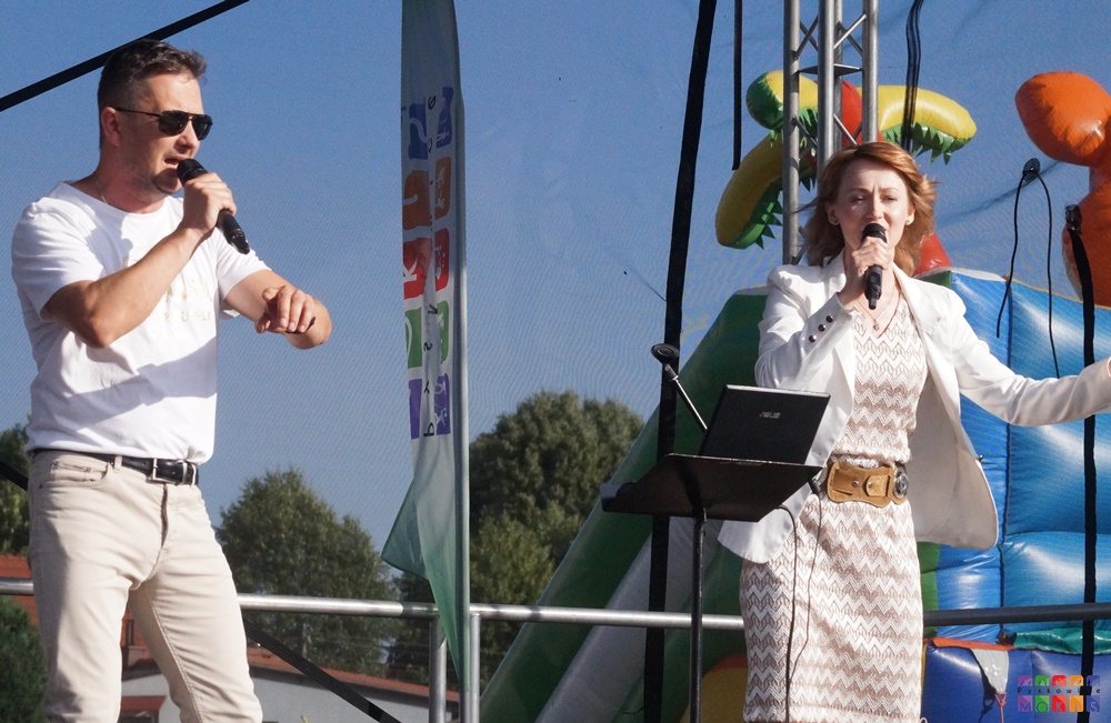 Zdjęcie przedstawia kobietę i mężczyznę z mikrofonem w ręku stojąca na scenie plenerowej. W tle powiewające flagi z logami.