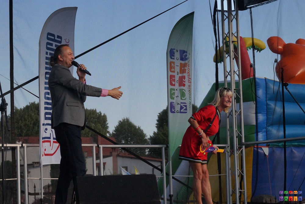 Zdjęcie przedstawia kobietę z zabawkową gitarą i mężczyznę z mikrofonem w ręku stojąca na scenie plenerowej. W tle powiewające flagi z logami.