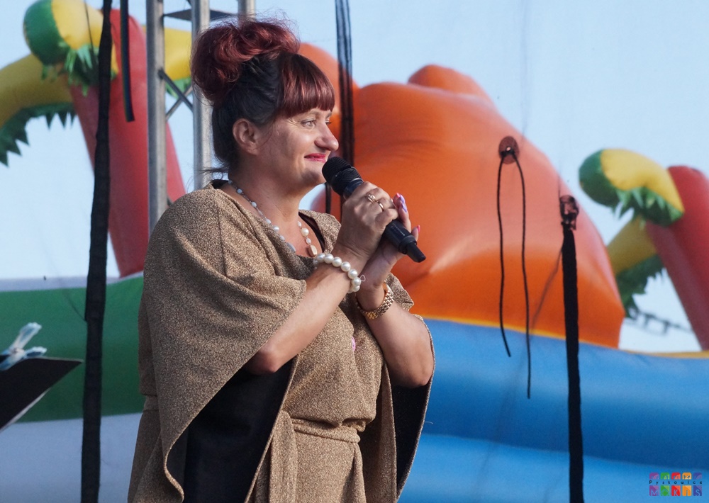 Zdjęcie przedstawia kobietę z mikrofonem w ręku stojąca na scenie plenerowej.