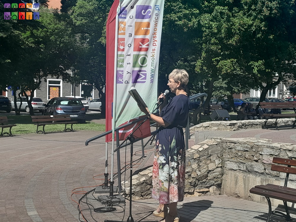 Zdjęcie przedstawia panią stojącą przy statywie mikrofonowym i trzymającą w lewej ręce otwartą książkę. W tle widać flagi oraz drzewa parkowe