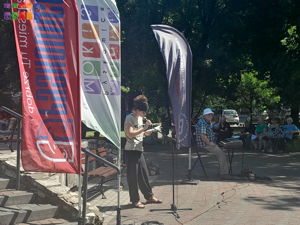 Zdjęcie przedstawia panią stojącą przy statywie mikrofonowym i trzymającą w lewej ręce otwartą książkę. W tle widać Pana siedzącego przy organach, flagi oraz drzewa parkowe