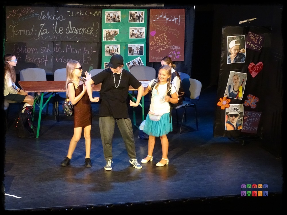 Zdjęcie przedstawia grupę aktorów na scenie w trakcie przedstawienia teatralnego. W tle scenografia - tablica lekcyjna z napisami, obrazki powieszone na zielonej ścianie.