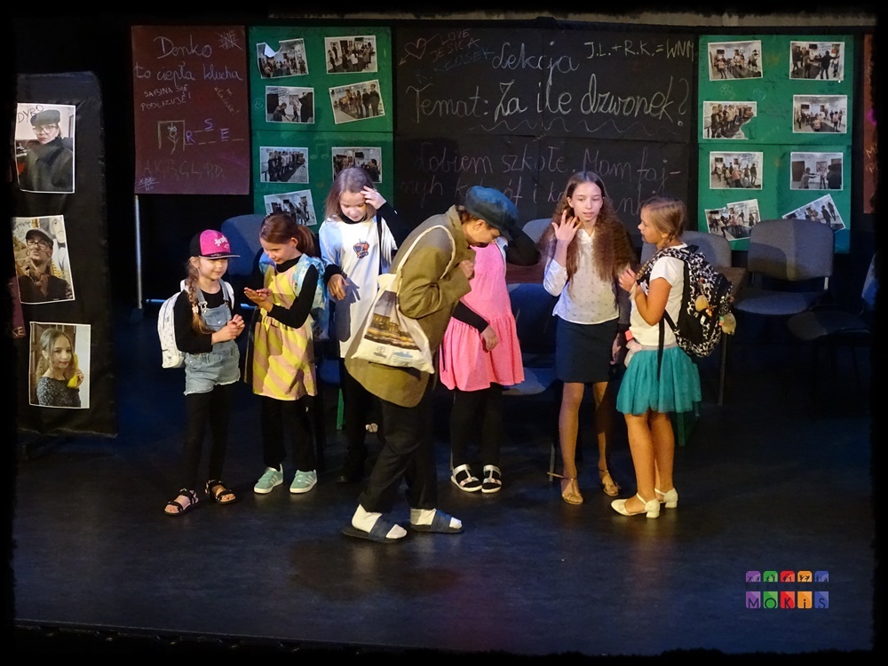 Zdjęcie przedstawia grupę aktorów na scenie w trakcie przedstawienia teatralnego. W tle scenografia - tablica lekcyjna z napisami, obrazki powieszone na zielonej ścianie.